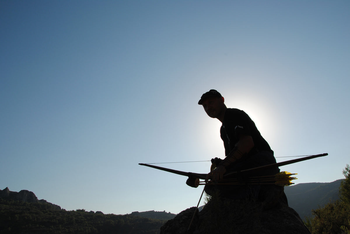 Im Vordergrund ist eine Silhouette eines knienden Menschen mit Mütze zu sehen, der einen Bogen in der Hand hält. Am Rücken hat er einen Köcher mit gelben Pfeilen. Der Himmel ist klar und blau, im Hintergrund sind Hügel und vereinzelte Büsche oder Bäume erkennbar. Die Sonne befindet sich direkt hinter dem Kopf der Person, wodurch ein starker Gegenlichteffekt entsteht. Die Szenerie wirkt ruhig und naturnah.