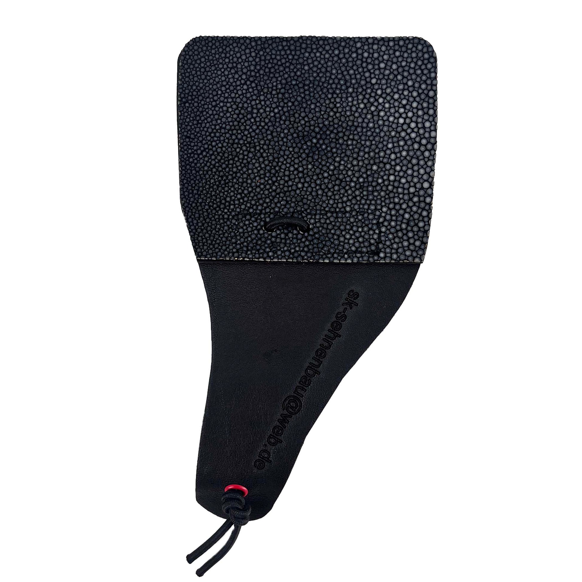 Ein flaches, schwarzfarbenes Lederobjekt, oben rechteckig mit Perlrochen-Struktur, unten glattes Leder mit geprägtem Text und schwarzer Kordel.