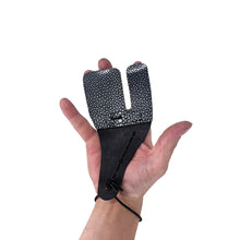 Eine Hand hält ein zweigeteiltes Tab aus Perlrochenleder, schwarzes genarbtes Material, schwarze Kordel um das Handgelenk.