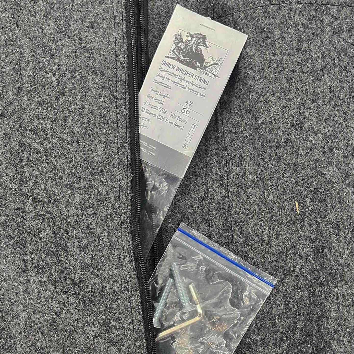 Grauer Filzstoff mit Reißverschluss, ein Beutel mit Schrauben und Innensechskantschlüssel sowie ein Etikett mit schwarzer Schrift und einer Zeichnung oben. Das Etikett beschreibt „Shrew Whisper String“ mit technischen Angaben.